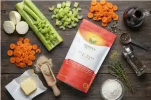 A top-down view of all ingredients for Beef Broth Gravy with Roasted Vegetables on a rustic wooden table. The Simply Halal Beef Bone Broth powder bag is centered, surrounded by chopped carrots, celery, onions, butter, flour, whole peppercorns, dried herbs, and a measuring spoon of broth powder, ready for preparation.