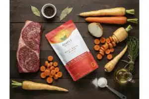A top-down flat lay displays all the fresh ingredients for Braised Beef with Root Vegetables on a rustic wooden background. A Simply Halal Beef Bone Broth powder bag is centered, surrounded by raw Halal beef brisket, whole and chopped carrots, parsnips, onion, bay leaves, black peppercorns, and olive oil, arranged for cooking.