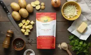 A flat lay of ingredients for Beef Broth Mashed Potatoes on a dark table. The Simply Halal Beef Bone Broth powder bag is centered, surrounded by cubed raw potatoes, diced onion, a cube of butter, black peppercorns, and fresh parsley, arranged for the recipe.