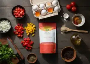 A flat lay of all fresh and dried ingredients needed for Halal Beef Broth Shakshuka. A Simply Halal Beef Bone Broth powder bag is centered, surrounded by tomatoes, diced onion and bell pepper, four eggs grouped at the top, cumin, paprika, and fresh parsley, all ready for cooking